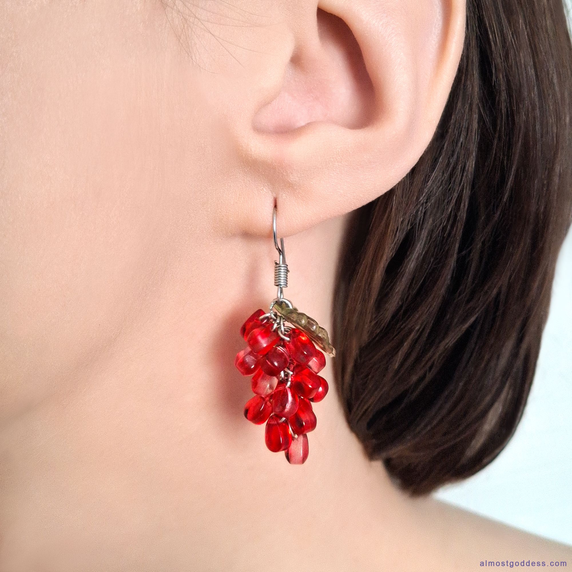 Red pomegranate seed dangle earrings with green acrylic leaf on stainless steel fishhook wire. Glass bead cluster earrings worn on ear, hypoallergenic fruit jewelry for sensitive ears