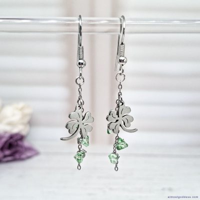 Stainless steel four-leaf clover drop earrings on acrylic stand featuring green glass beads on chains. Hypoallergenic handmade dangle earrings for women