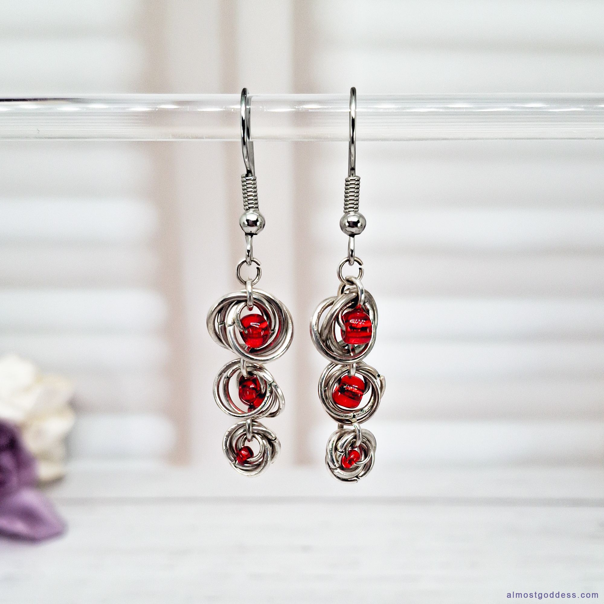 Pair of silver chainmaille dangle earrings featuring three tiers of linked metal rings with small faceted red beads in the center, hanging from fishhook ear wires.